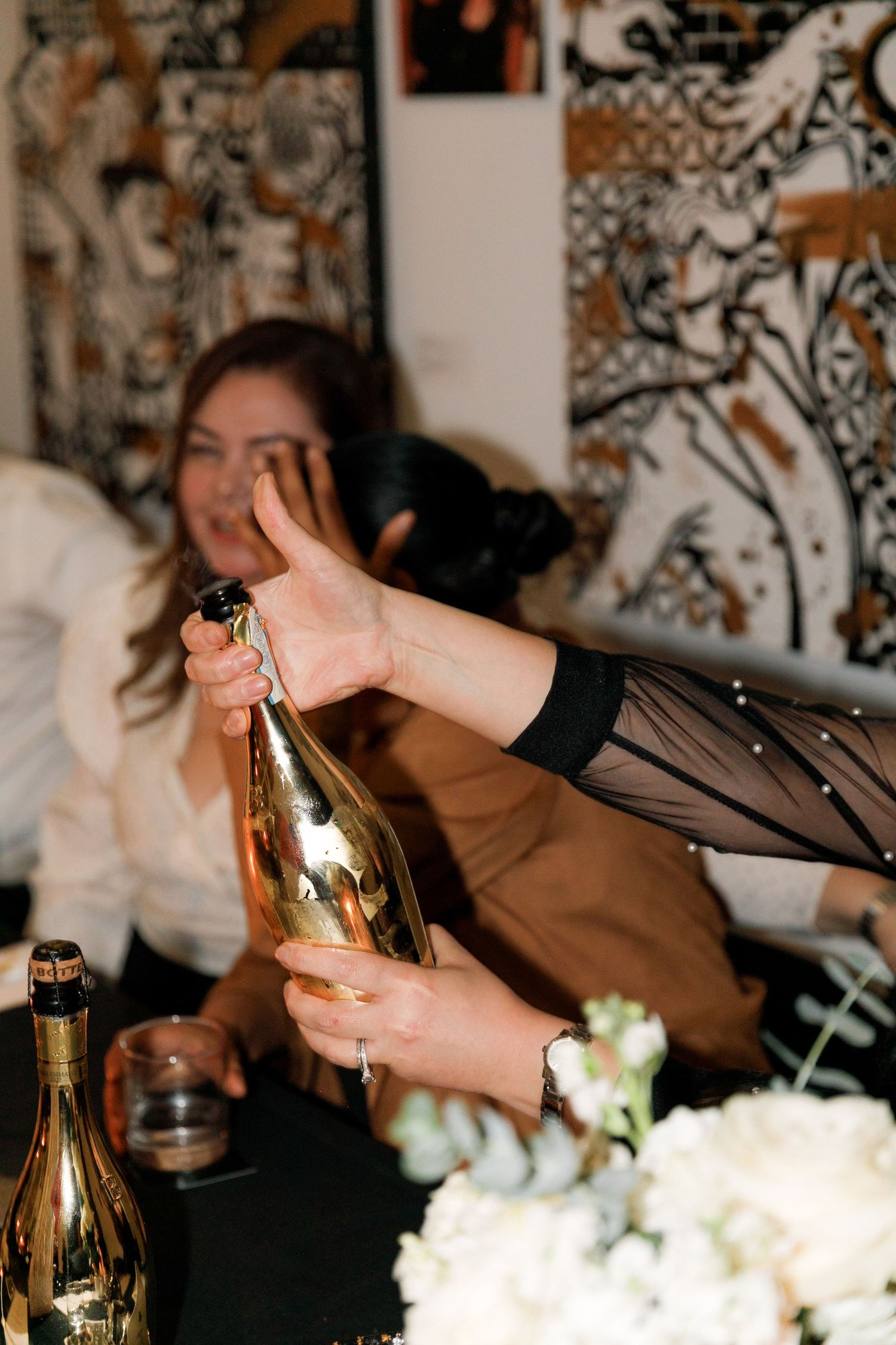 Hands holding a gold champagne bottle at a celebration party with guests in the background.
