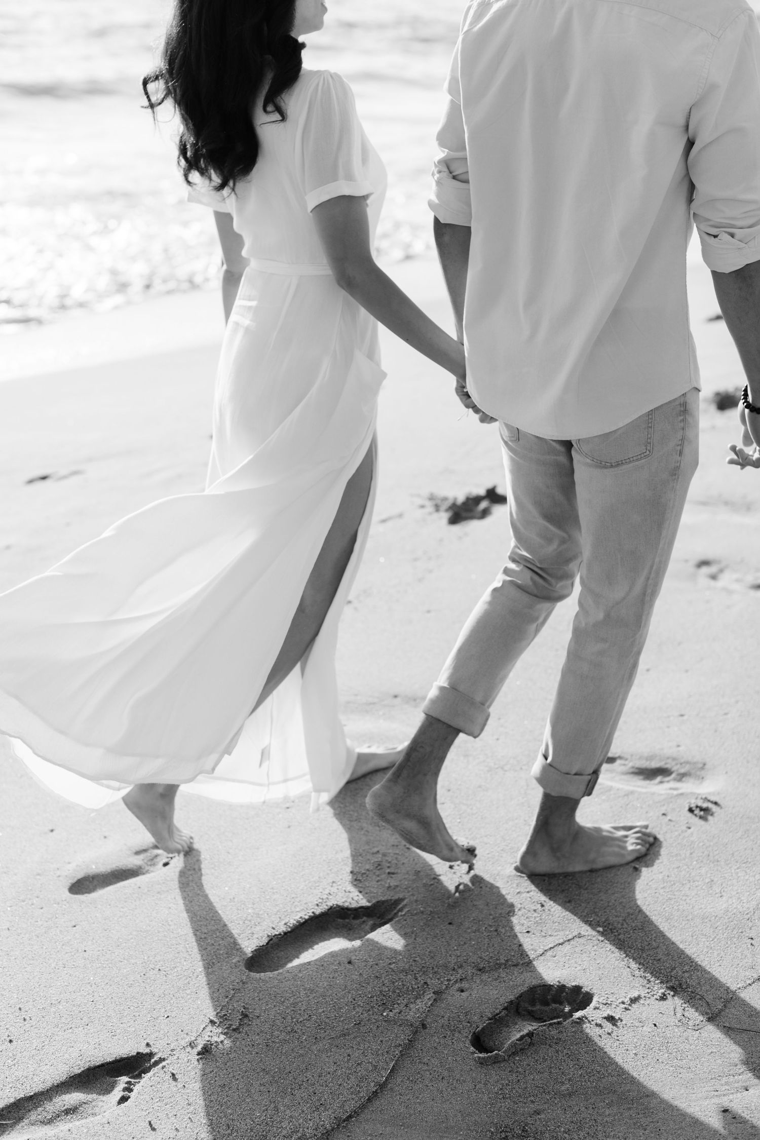 Black and white photo of a couple holding hands walking barefoot on a sandy beach leaving footprints.