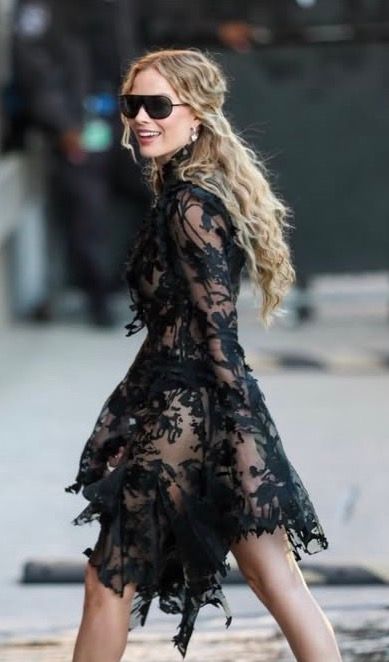 Woman in sheer black lace dress with long sleeves, wearing sunglasses, smiling over shoulder, with curly blonde hair.