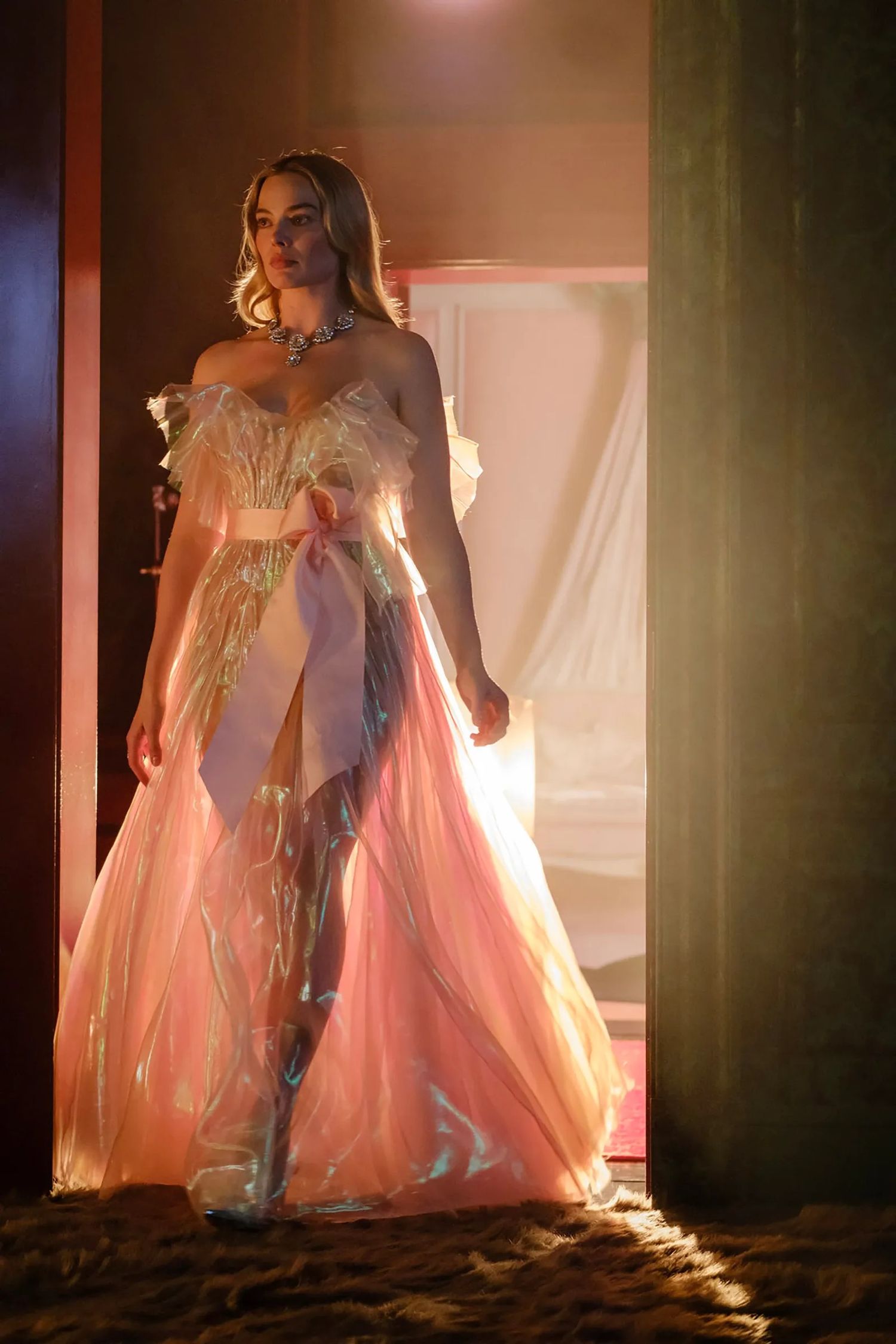 Woman in elegant pink tulle gown with sheer overlay and statement necklace poses dramatically in warm golden light.