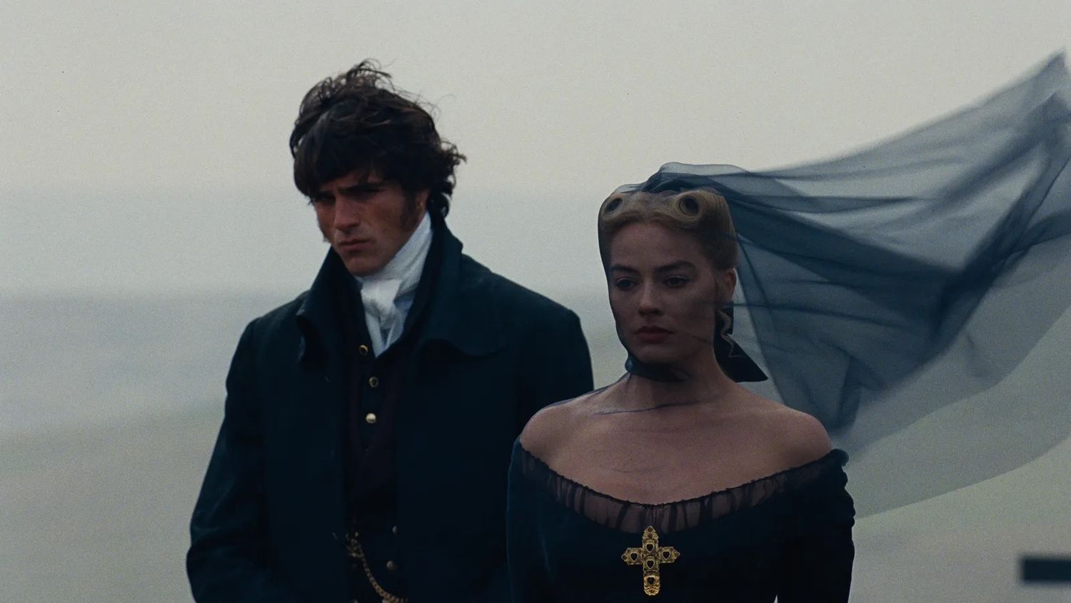 A man and woman in Victorian period costumes stand outdoors in misty fog, woman wearing black dress with flowing blue veil.