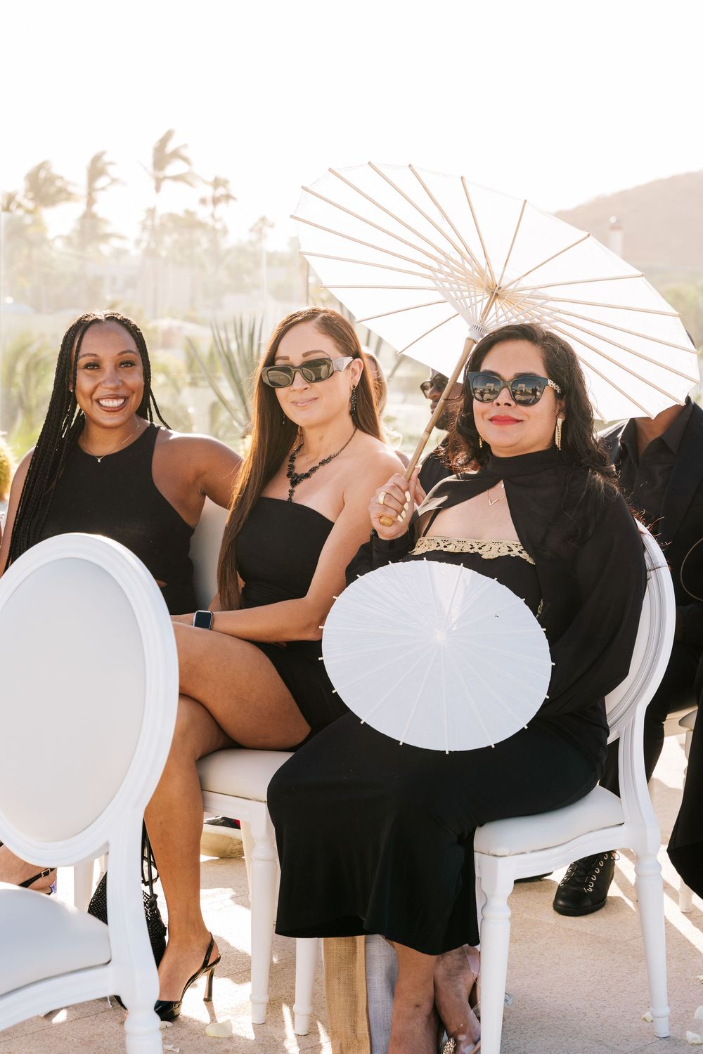 Three women in black dresses holding white parasols seated in white chairs at an outdoor wedding ceremony.