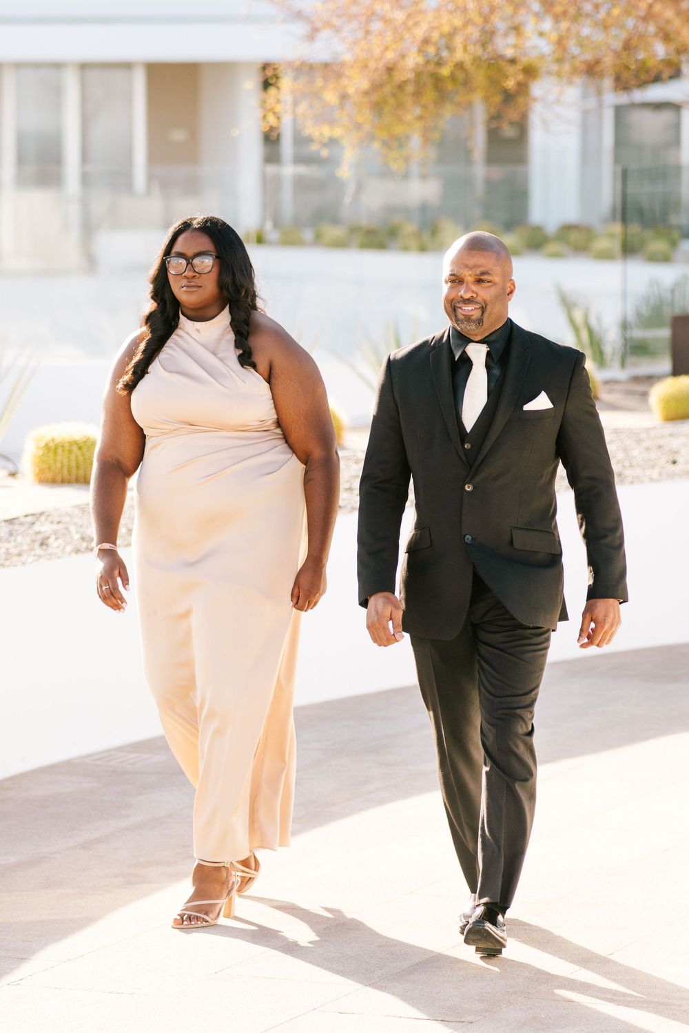 Elegant couple walking outdoors, woman in champagne halter gown and man in black tuxedo, in a sunny modern setting.