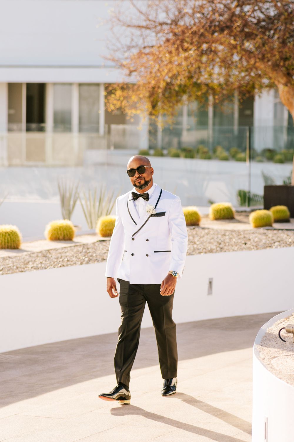 Stylish groom in white double-breasted tuxedo jacket, black bow tie, and sunglasses posing outdoors at wedding venue.