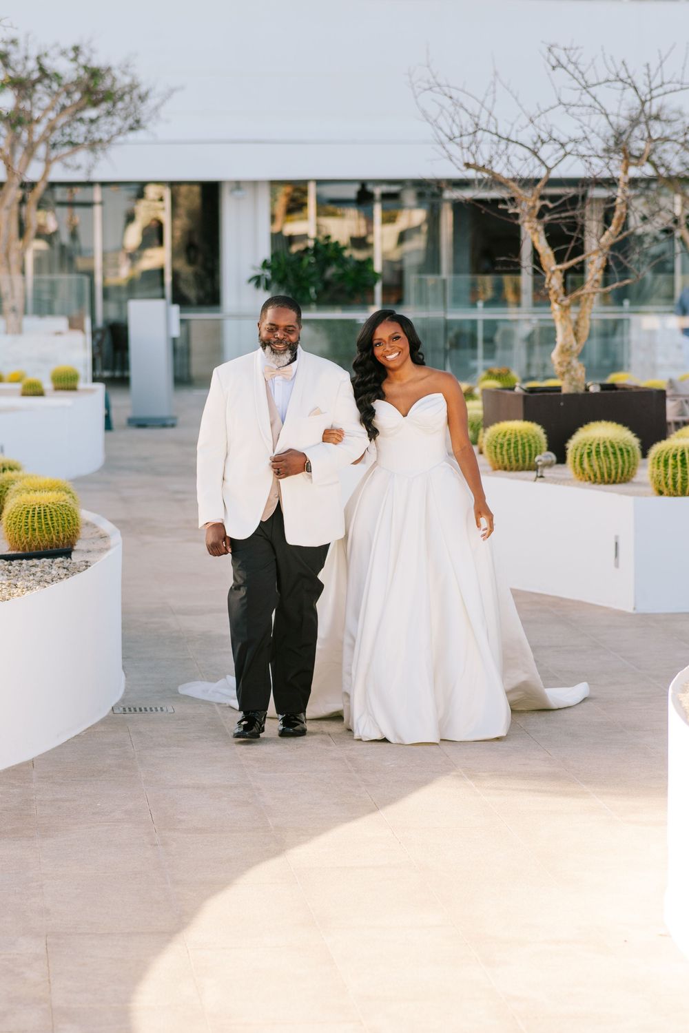 Bride in strapless white gown walks arm-in-arm with groom in white jacket at a modern resort venue.