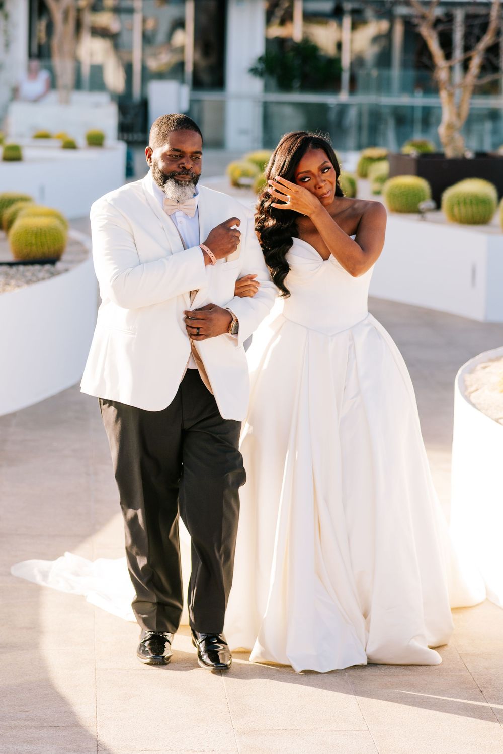 Emotional bride in strapless white gown wipes tears beside groom in white tuxedo jacket during outdoor wedding.