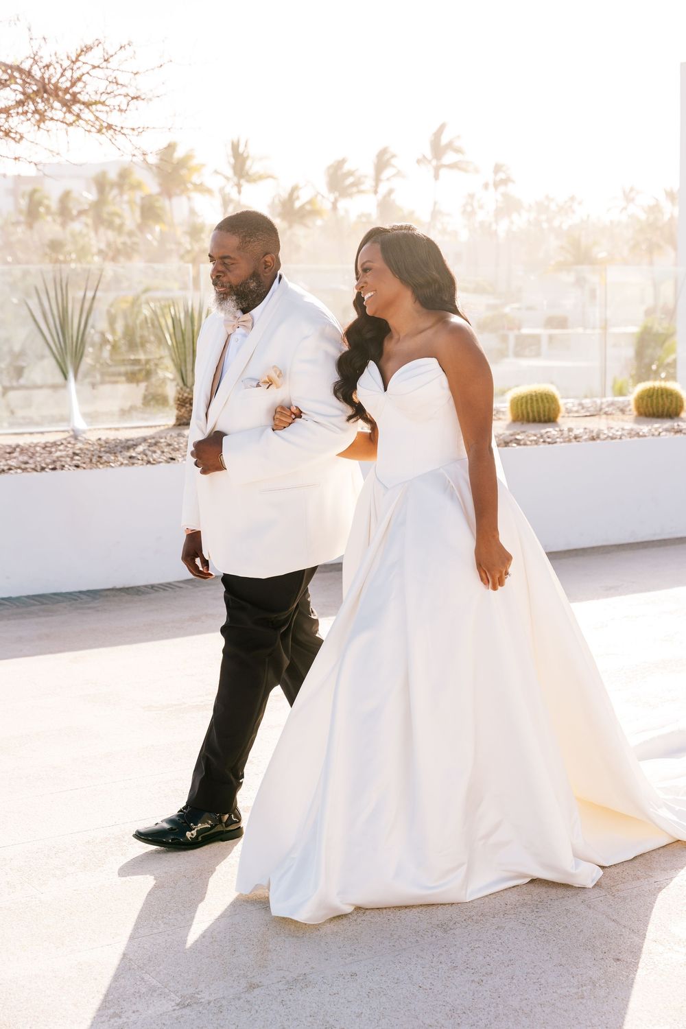 Bride in elegant strapless ball gown walks with groom in white tuxedo at sunny outdoor wedding venue.