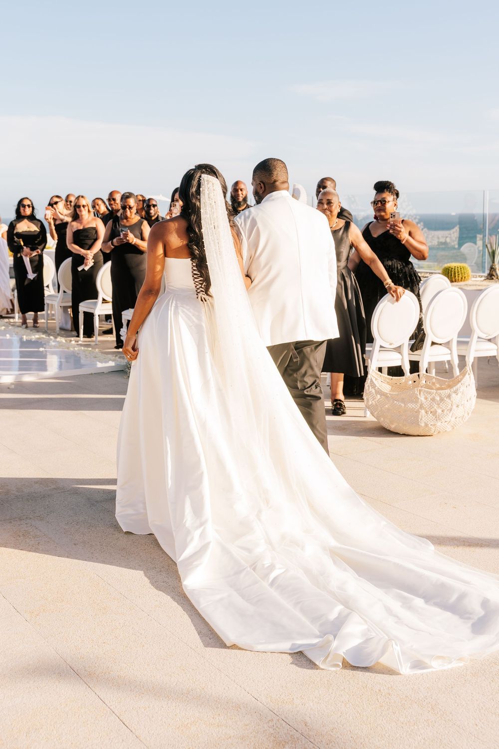 Bride in white gown with long veil walks toward wedding guests dressed in black at outdoor rooftop ceremony.