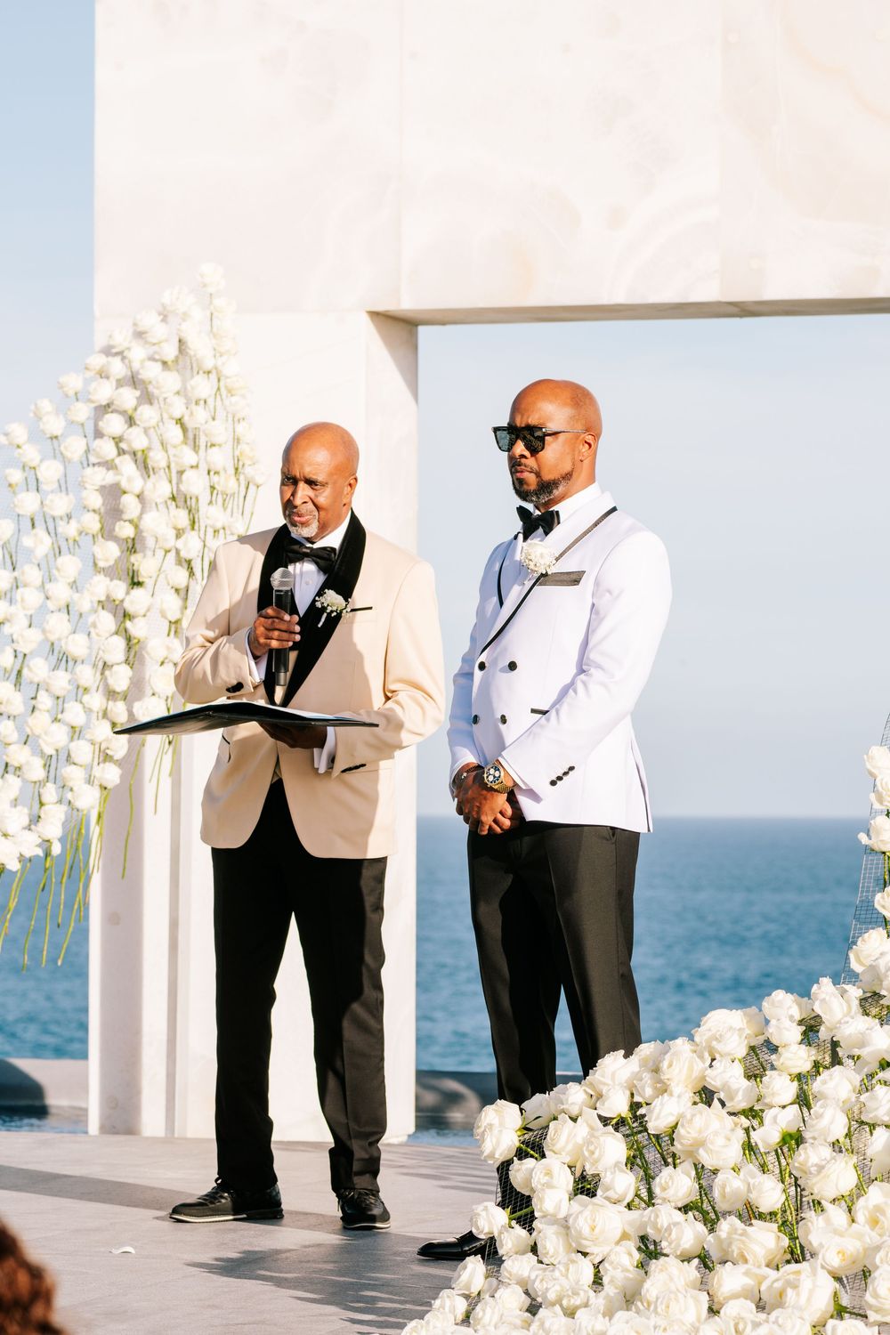 Officiant in beige tuxedo reads vows beside groom in white tuxedo at elegant outdoor wedding with white floral backdrop.