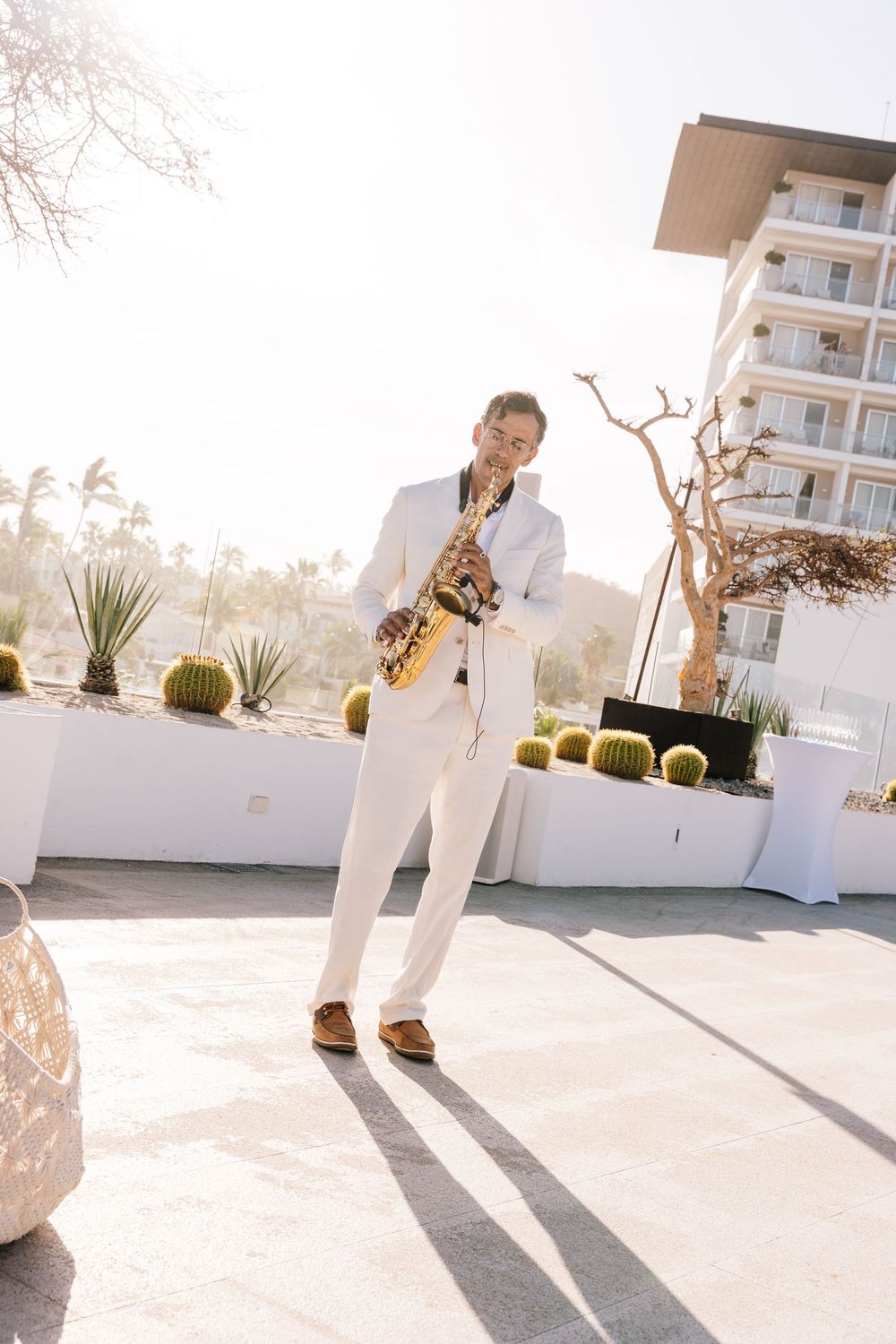 Elegant saxophonist in white suit performing outdoors at a sunny resort with cacti and palm trees.