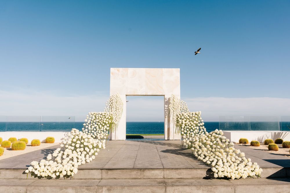 White floral arch with cascading roses frames ocean view under clear blue sky at elegant outdoor wedding ceremony.