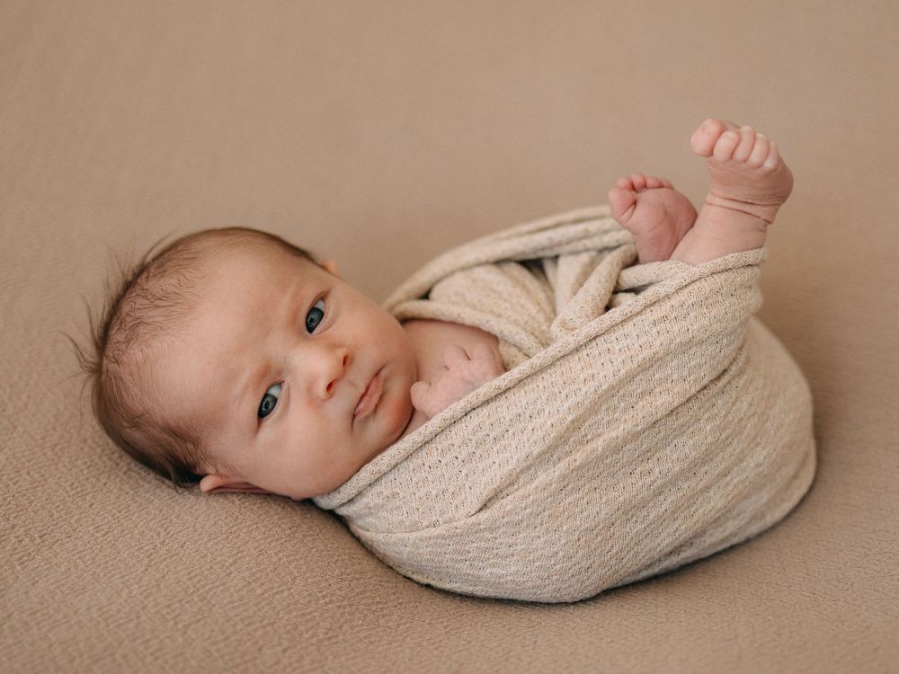 Newborn baby swaddled in a cream knit wrap, lying on a beige blanket with one tiny fist raised.