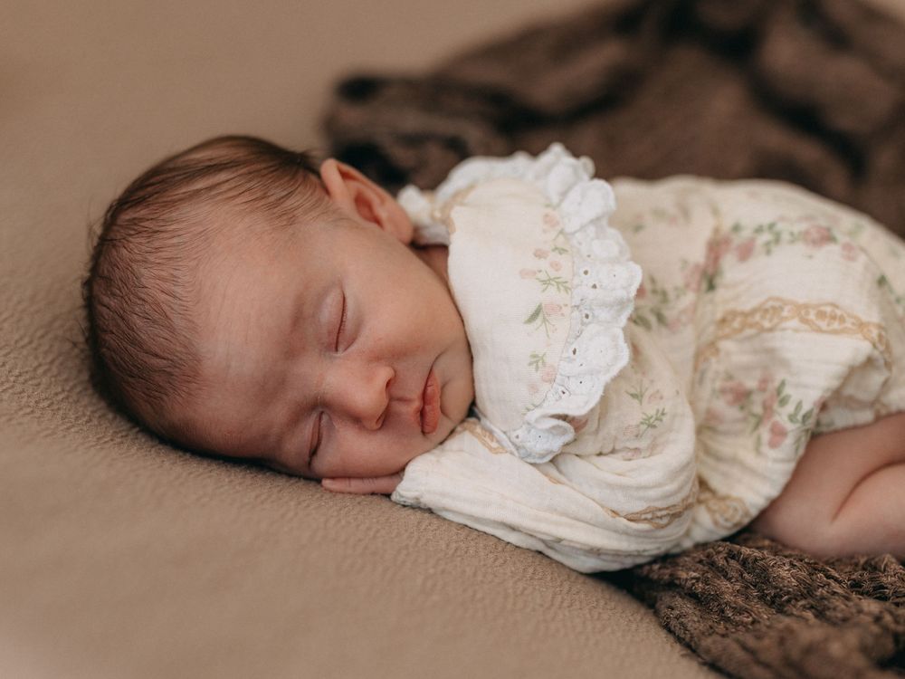 Peaceful newborn baby sleeping in a white floral outfit on a soft beige blanket.