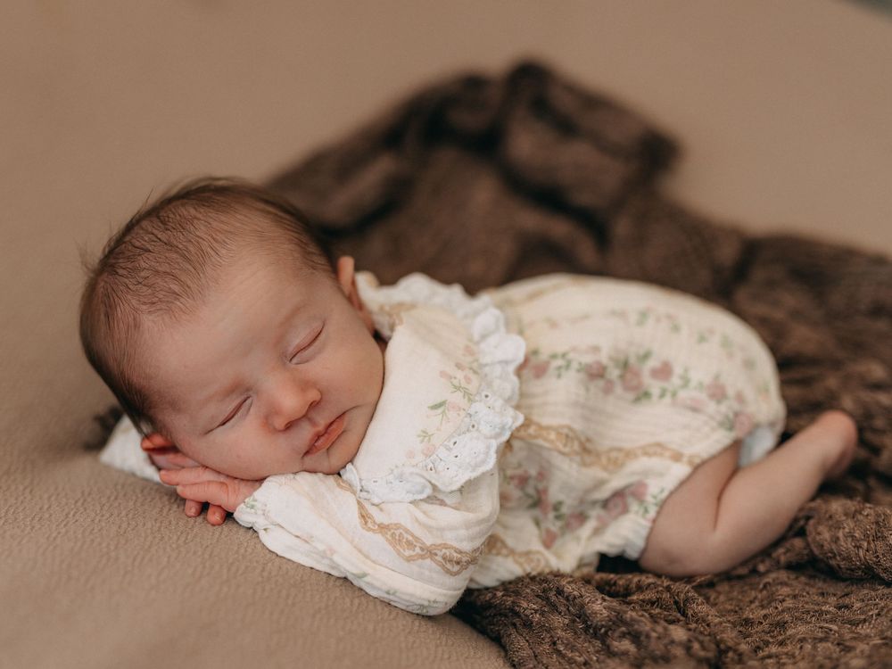 Peaceful newborn baby sleeping in a floral romper on a soft brown blanket in a professional photo session.