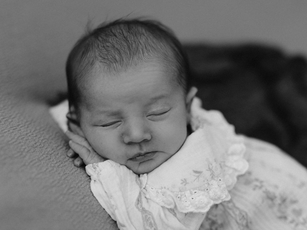 Black and white newborn baby sleeping peacefully in a white lace outfit, resting cheek on tiny hands.