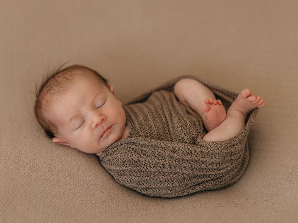 Peaceful newborn baby swaddled in a brown knit wrap, sleeping on a tan backdrop.