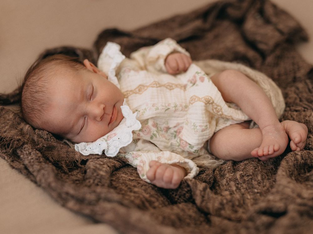 Peaceful newborn baby sleeping on a brown knit blanket, wearing a delicate white lace romper.