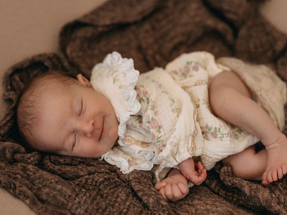 Sleeping newborn baby in vintage floral lace dress on brown knit blanket.