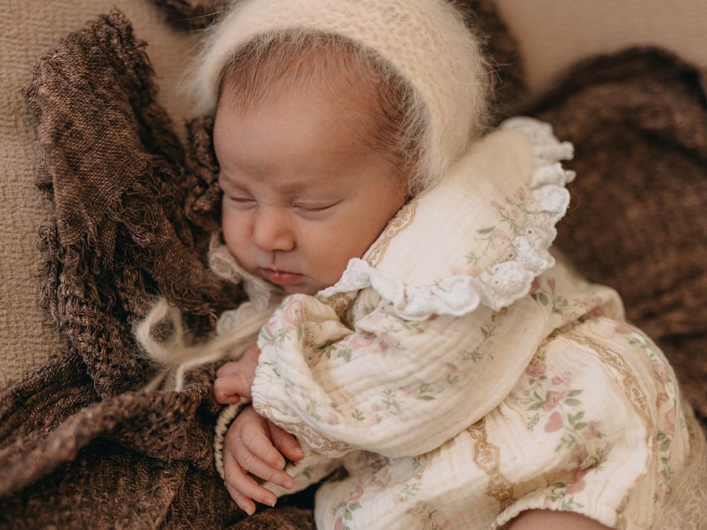 Sleeping newborn baby in floral romper and knit bonnet on brown textured blanket.