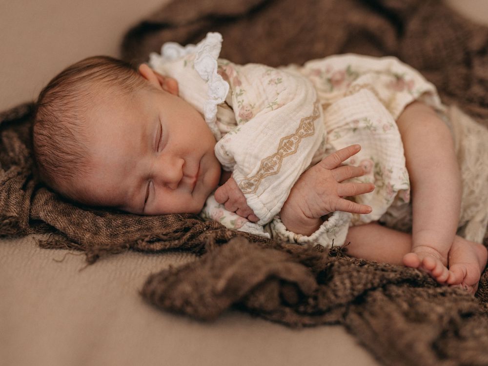 Peaceful newborn baby sleeping in a vintage floral outfit on a brown knit blanket in a professional photo.