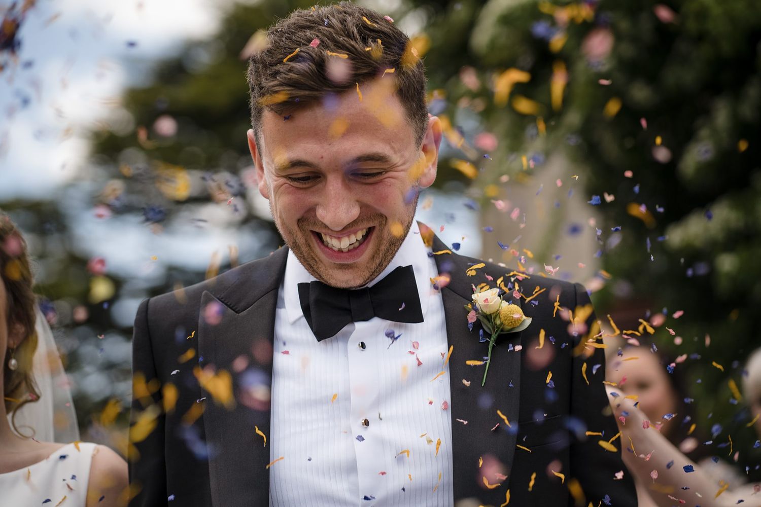 Joyful groom in black tuxedo smiling as colorful confetti rains down during outdoor wedding celebration.
