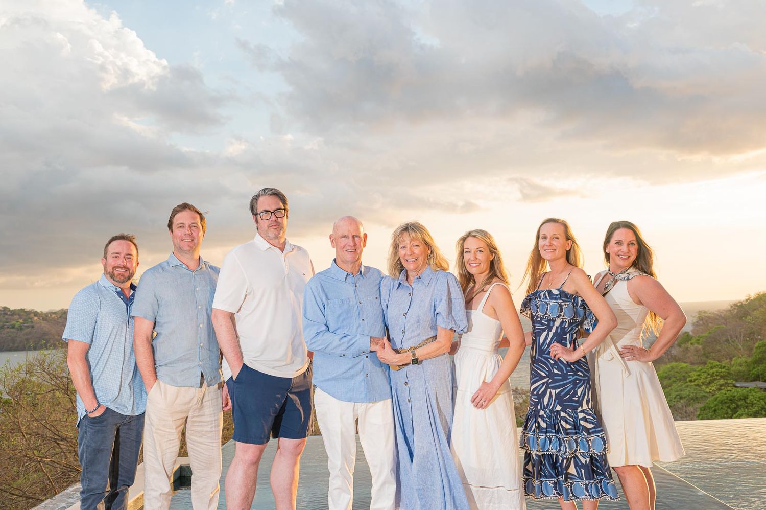 A joyful family group of seven poses together outdoors at sunset with a scenic tropical landscape backdrop.