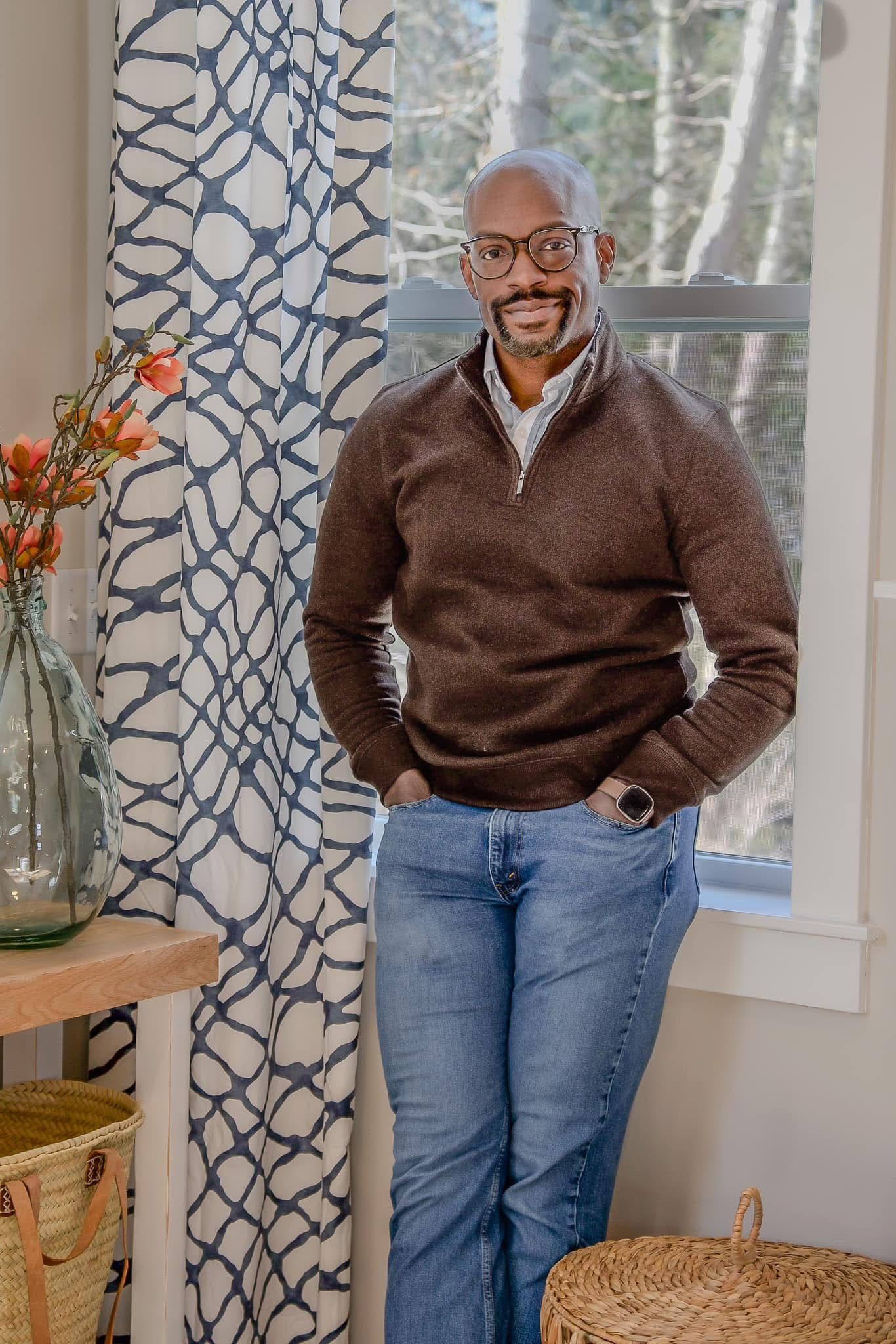 Smiling man in brown quarter-zip sweater and jeans stands by window with patterned curtains in stylish home interior.