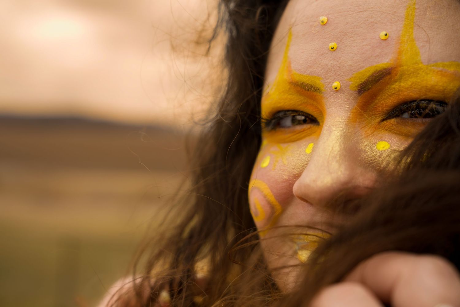Close-up portrait of a woman with vibrant yellow face paint and gold dot embellishments against a blurred outdoor backdrop.