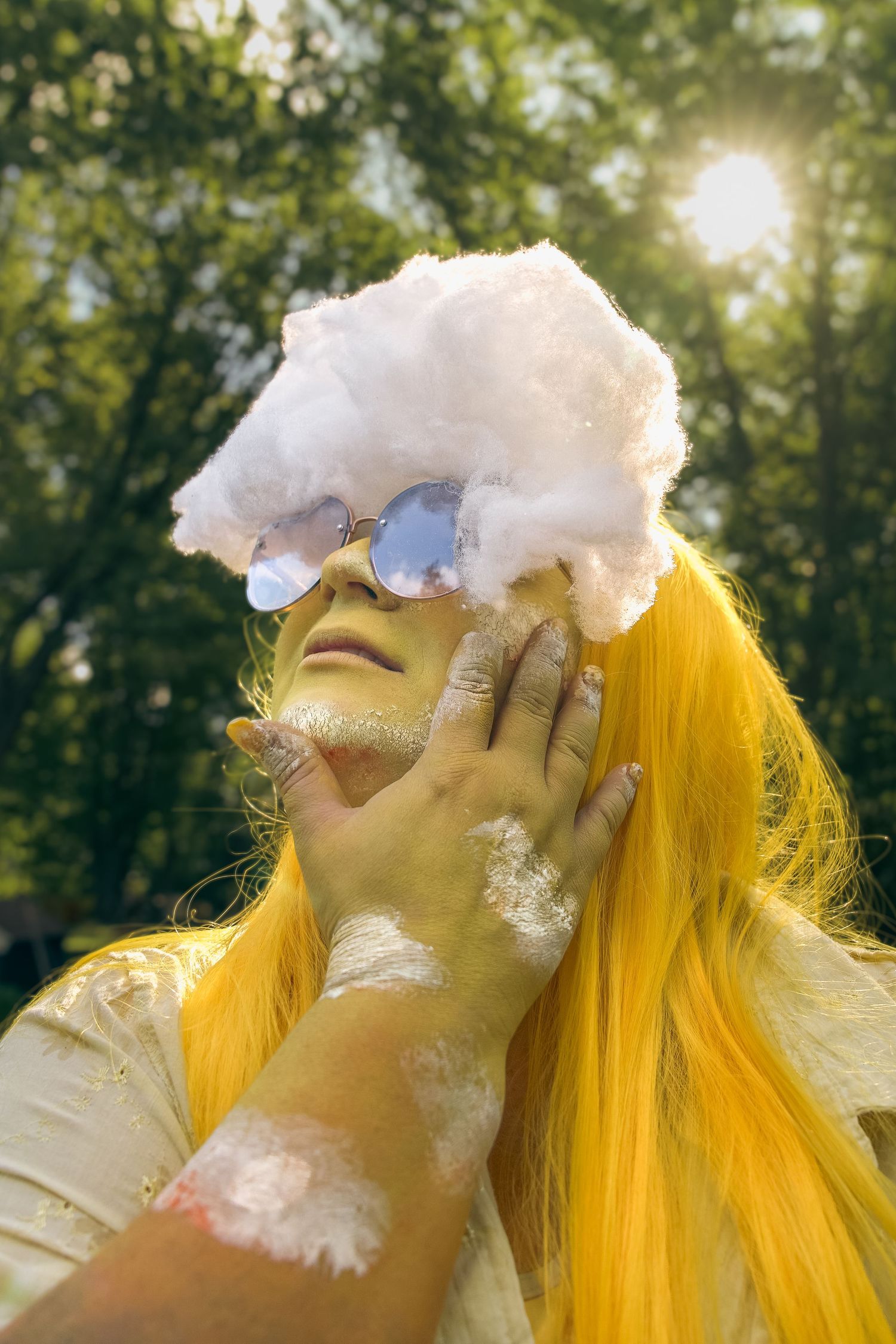 Person with yellow hair, blue sunglasses, and white powder, holding cotton candy on head outdoors in sunlight.