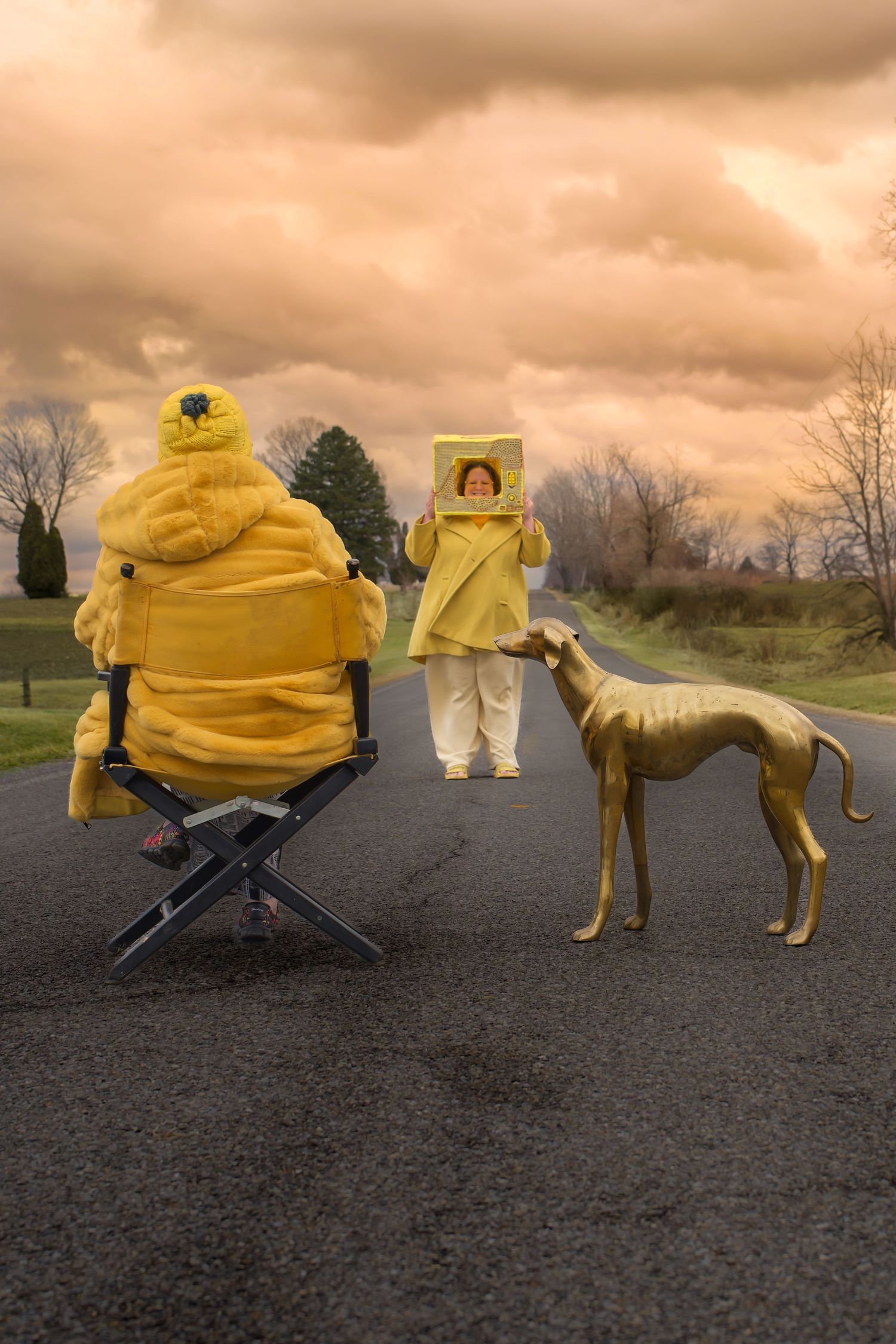 Person in yellow costume sits in director's chair as golden greyhound and woman holding box pose on road under dramatic sky.