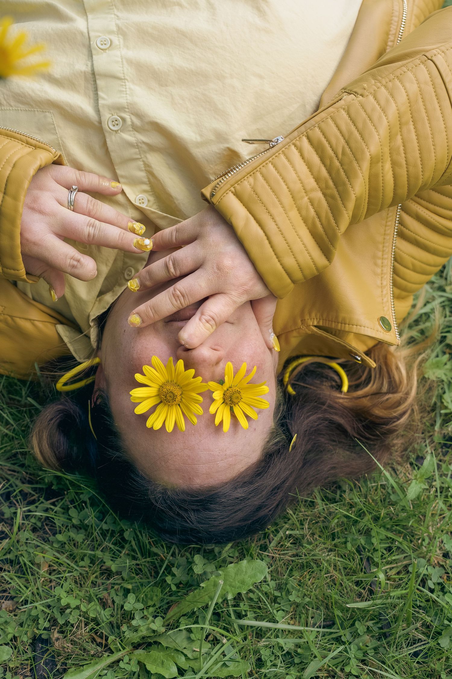 Woman lying on grass in yellow jacket with yellow daisies placed over her eyes, hand raised near face.