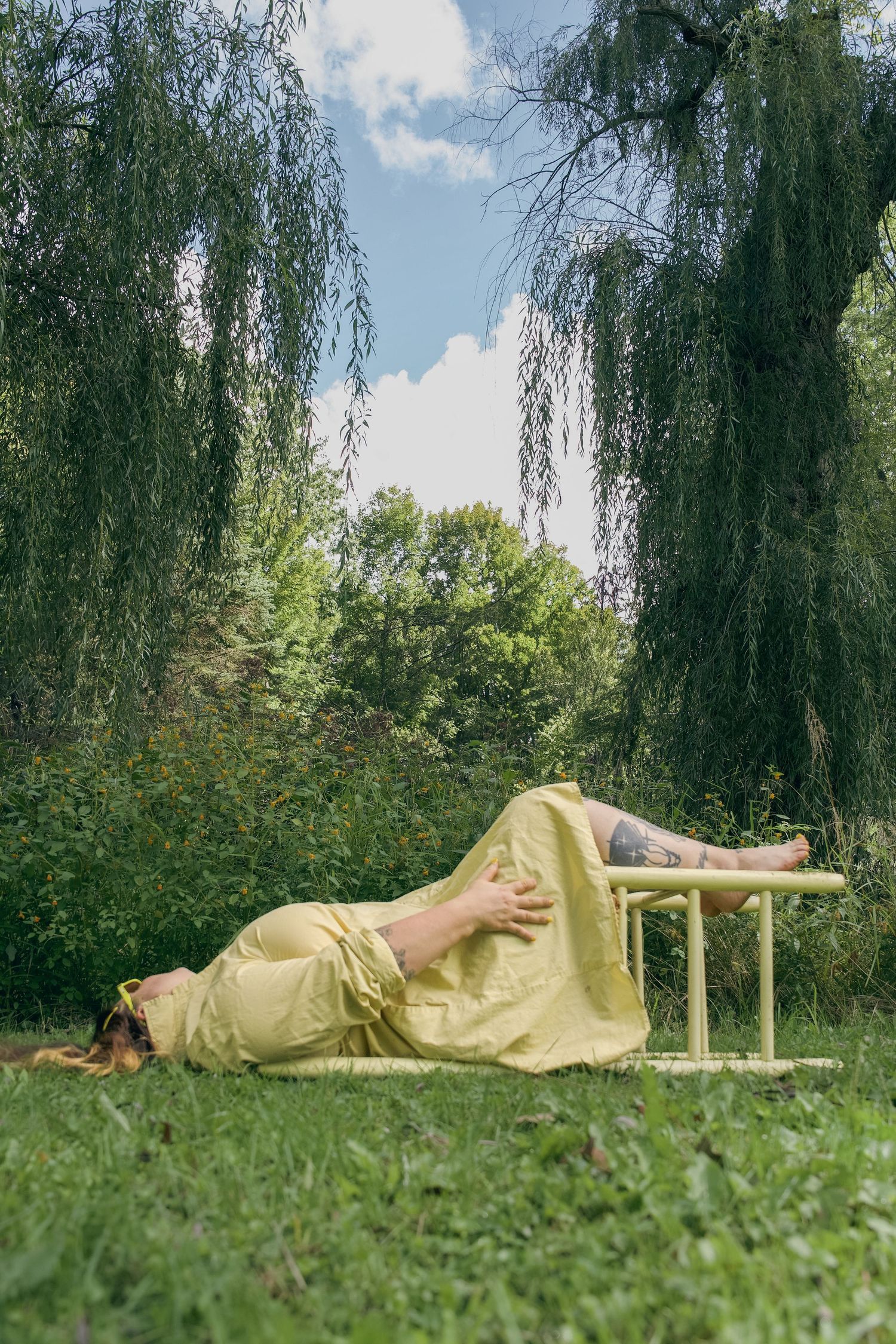Person in yellow dress lying on bench under weeping willow trees in lush green garden on sunny day.
