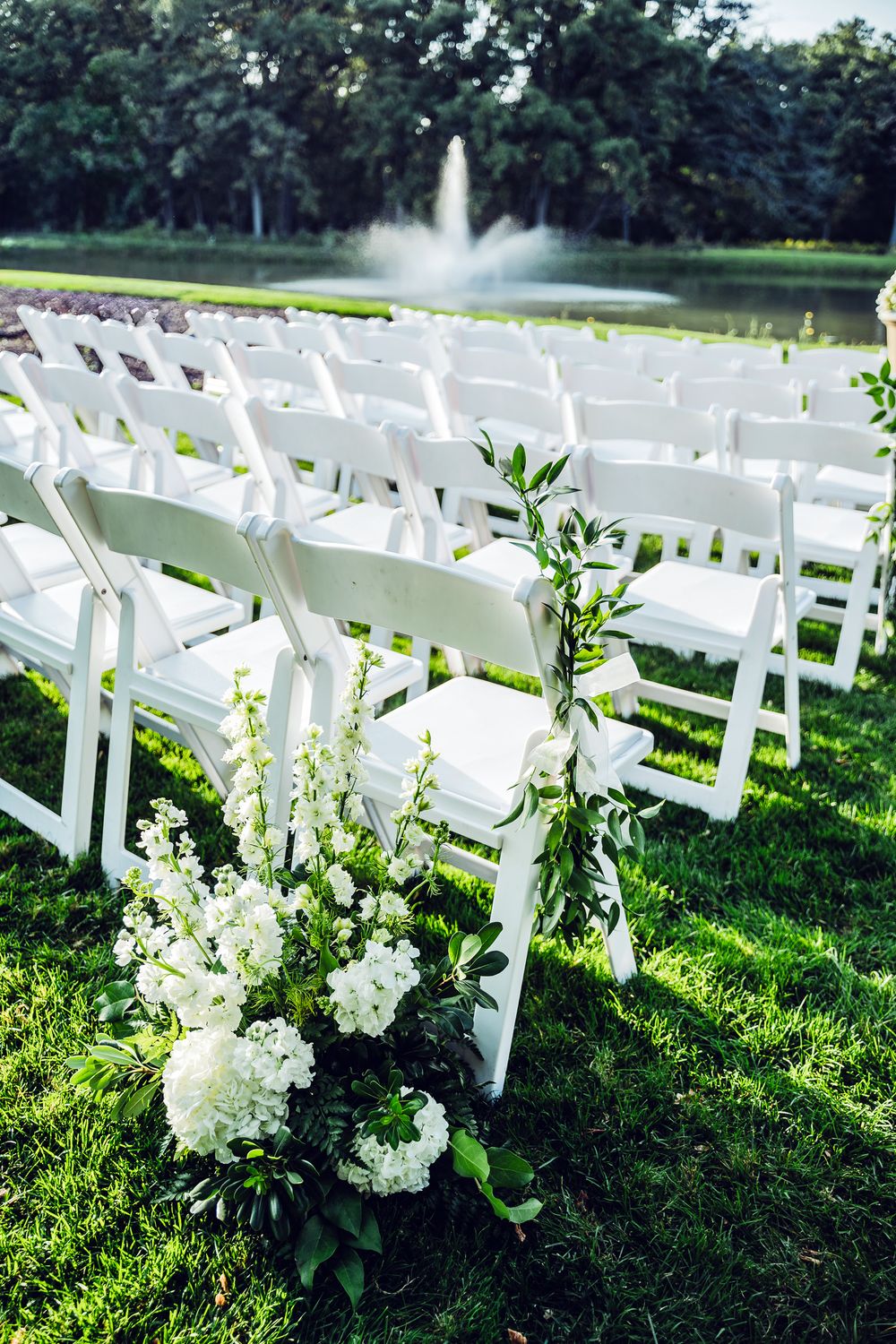 White folding chairs arranged in rows on green grass with white floral arrangements and a fountain backdrop for an outdoor wedding.