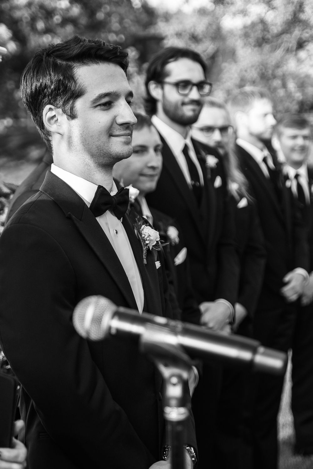 Groom in black tuxedo smiling at altar with groomsmen standing in line behind him at outdoor wedding ceremony.