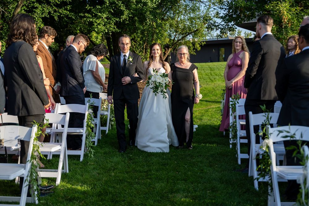 Bride and groom walk down outdoor wedding aisle flanked by guests seated in white chairs on lush green lawn.