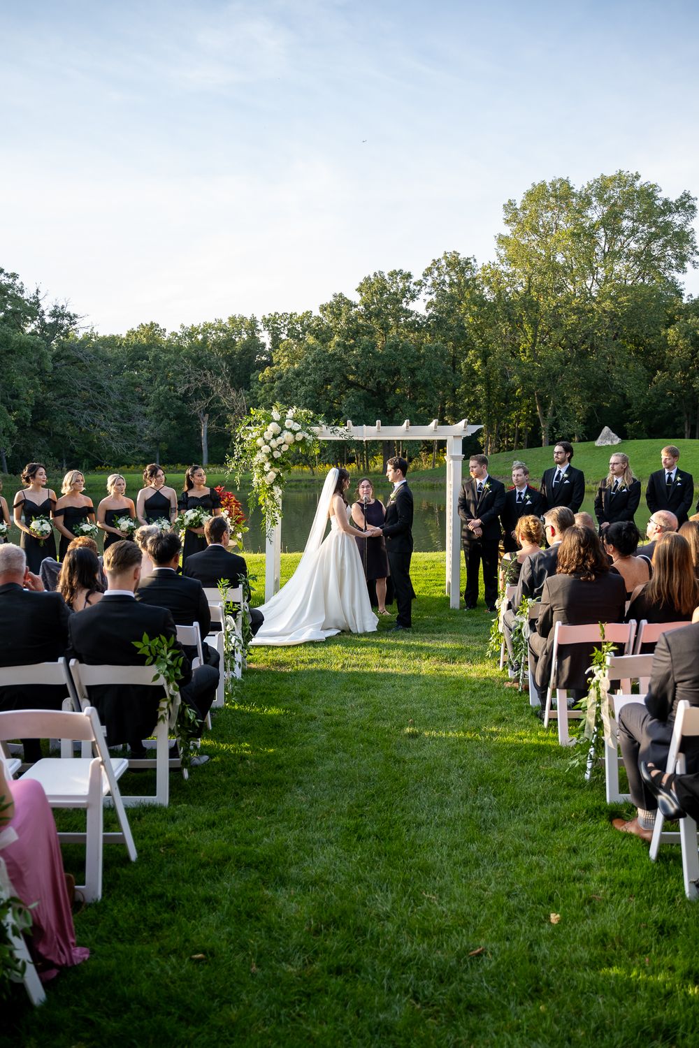Outdoor wedding ceremony on green lawn with white chairs, floral arch, bride and groom at altar, bridesmaids in black dresses.