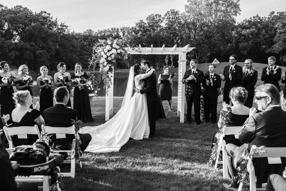 Black and white outdoor wedding ceremony with bride and groom kissing at floral arch, guests seated on white chairs.