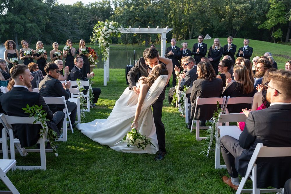 Bride and groom share first kiss at outdoor wedding ceremony with white chairs and floral arch.