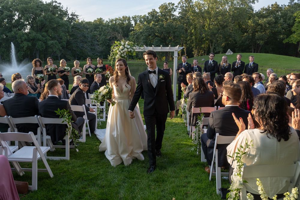 Bride and groom walk down outdoor wedding aisle surrounded by seated guests on a lush green lawn.
