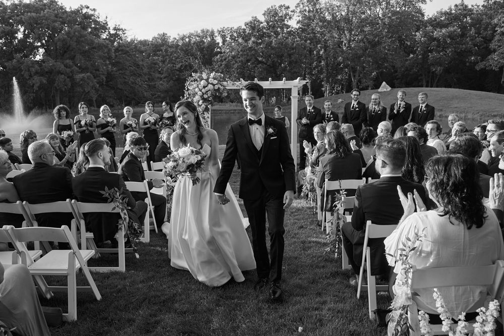 Black and white photo of a bride and groom walking down the aisle at an outdoor wedding ceremony.