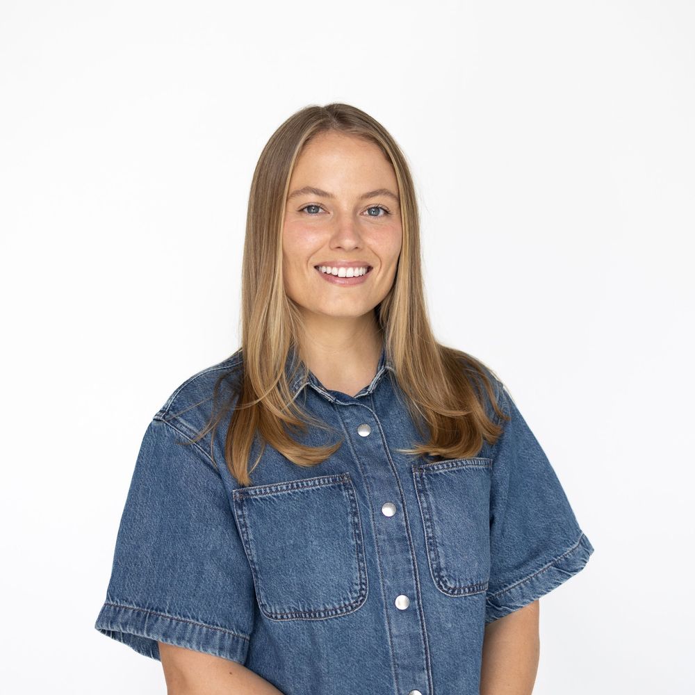 Smiling young woman with long blonde hair wearing a blue denim short-sleeve shirt against a white background.
