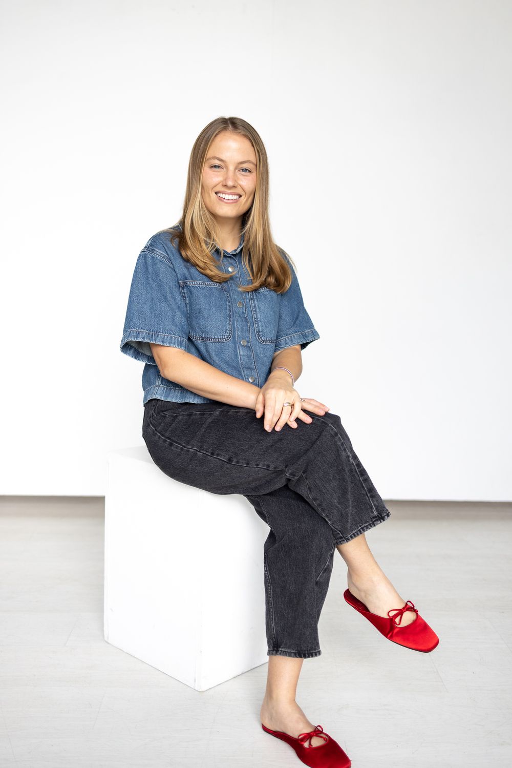 Smiling woman sitting on white cube wearing denim shirt, black jeans, and red flats against white background.