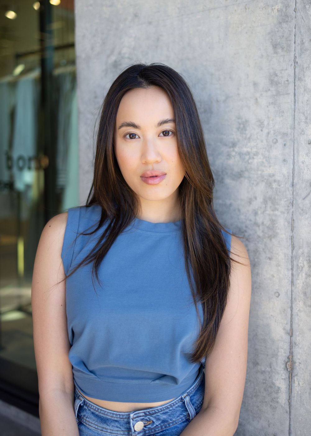 Professional portrait of someone in a blue sleeveless top and jeans against a concrete wall.