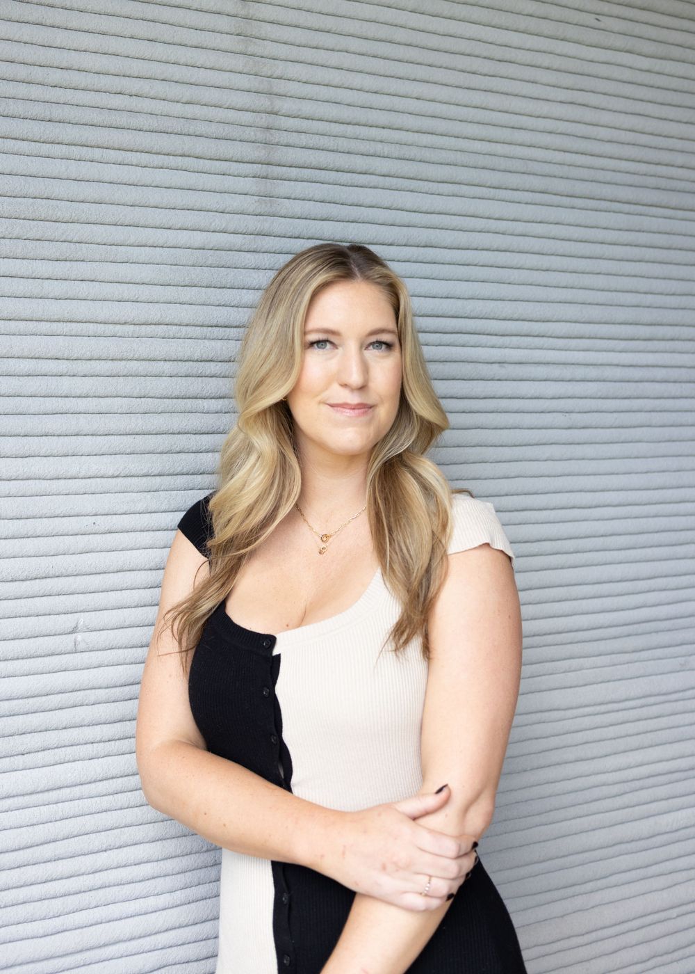 A confident blonde woman in a black and white outfit poses against a gray textured wall.