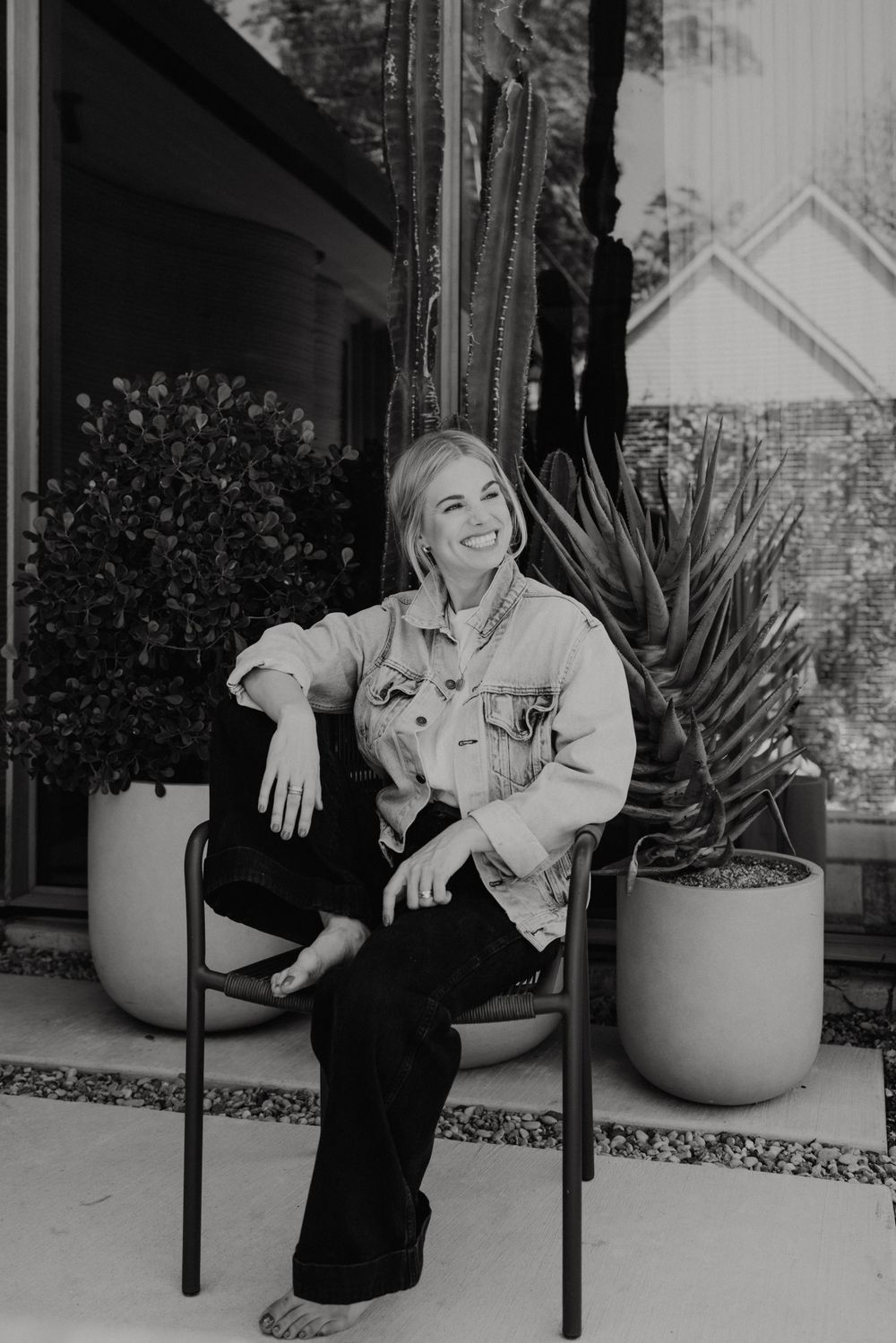 Black and white portrait of person sitting casually outdoors near potted plants.