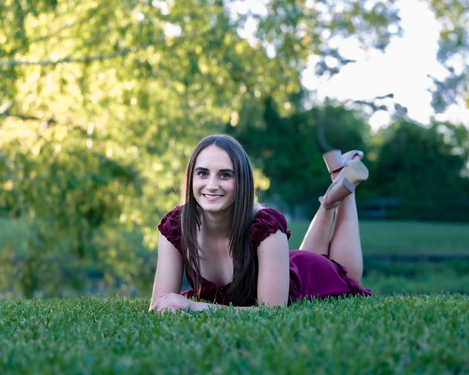 Young woman in burgundy dress smiling on a wooden swing in a lush green park on a sunny day.