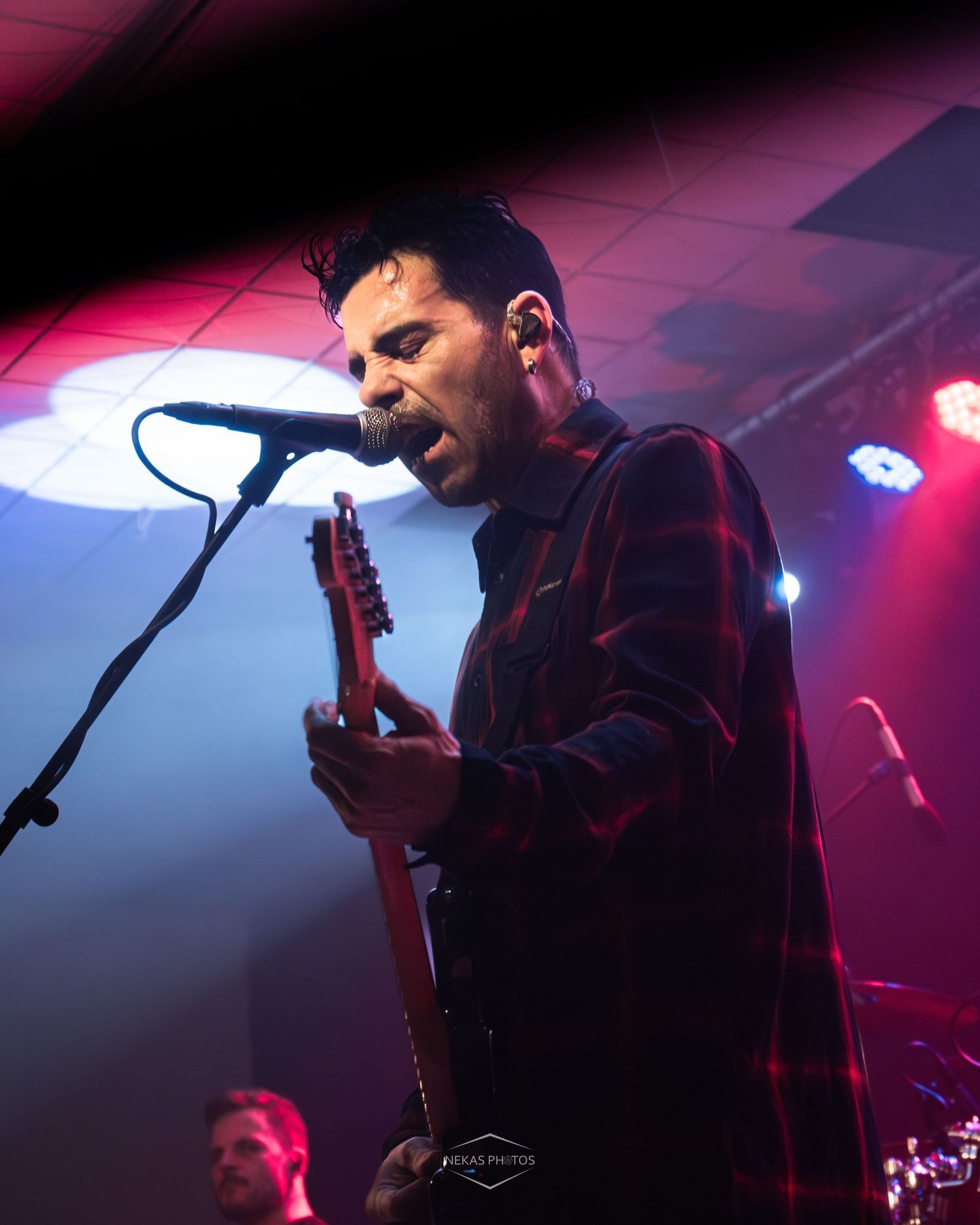 Male guitarist singing into microphone on stage under red and blue lights during live rock concert performance.