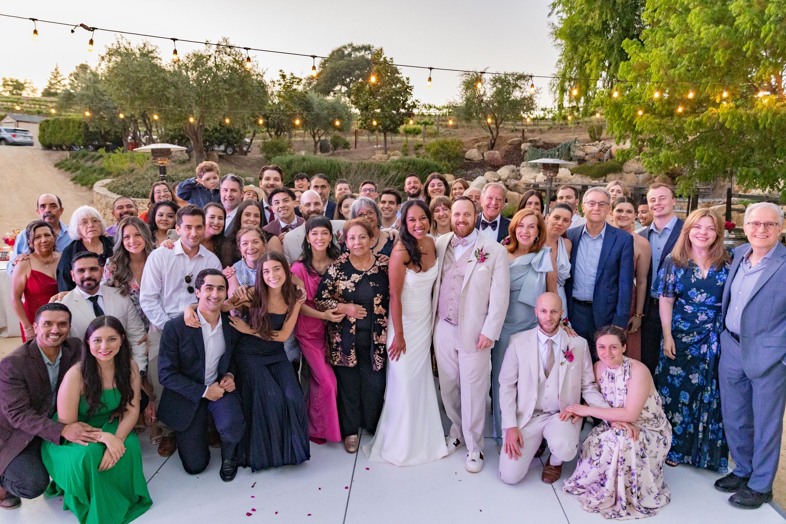 Large wedding party group photo with guests in formal attire on outdoor patio.