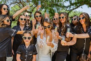 Celebratory toast with wedding party wearing matching black outfits and sunglasses in outdoor setting.