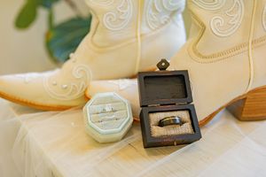 Wedding rings and ring boxes displayed next to elegant white cowboy boots with embroidered details.